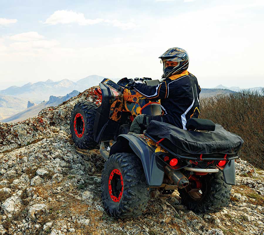 A man is riding a rugged, new-looking quad, and is poised atop a mountain overlooking a grand nature scene. He is looking over his shoulder at the camera, proud of the purchase he has financed using an LMG Financing dealership.