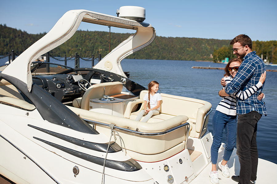 A white man and woman, each with brown hair, are hugging on a large, luxurious boat. A young child with a braid is smiling into the distance. They are on a lake, and it is a bright summer's day.