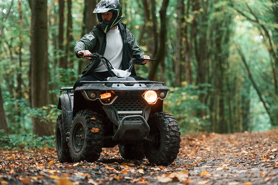 A man is riding a quad in a forest, leaves on the path around him. He is mid-action, looking off to the side of the frame.