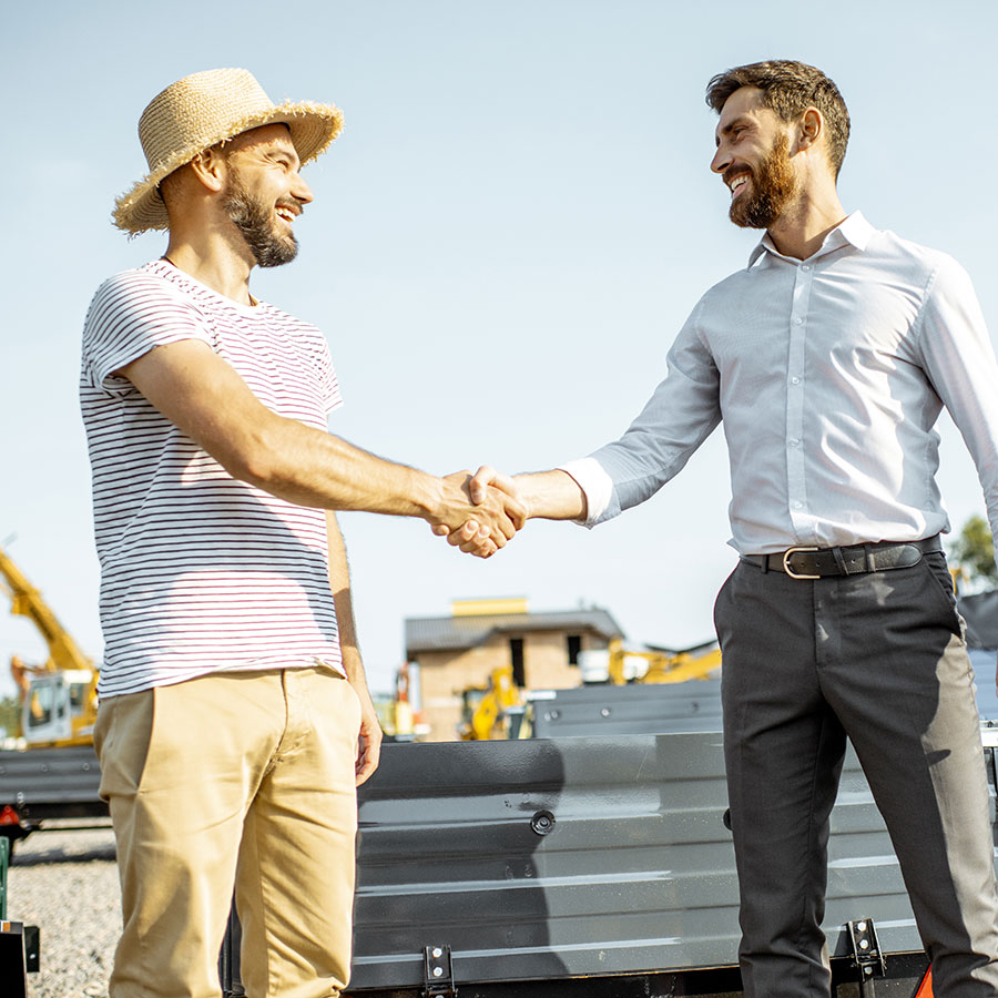 Two men shake hands in a sunny powersports yard. One is wearing a hat and the other is wearing a dress shirt and pants.