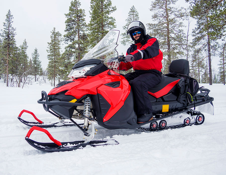 A man is wearing a red and black winter jacket, on a red and black snowmobile, smiling at the camera in a winter wonderland.
