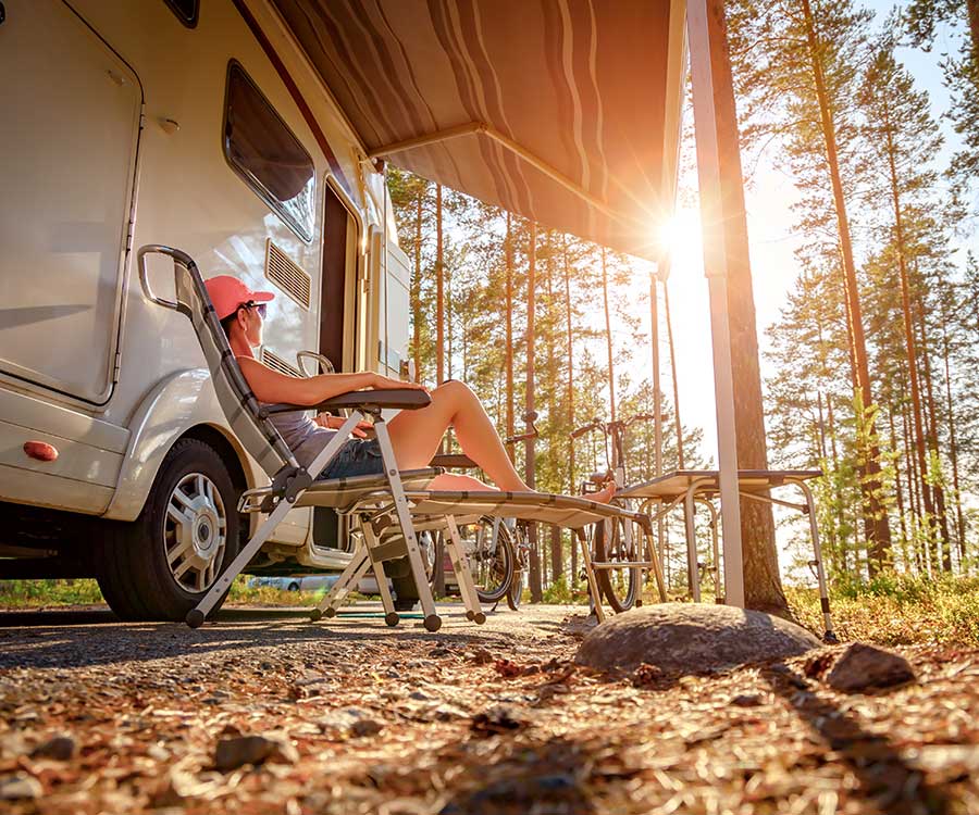 A white woman with a pink ballcamp relaxes on a camp chair in the late afternoon sun. She has her feet up and is under an awning alongsider her newly financed RV.