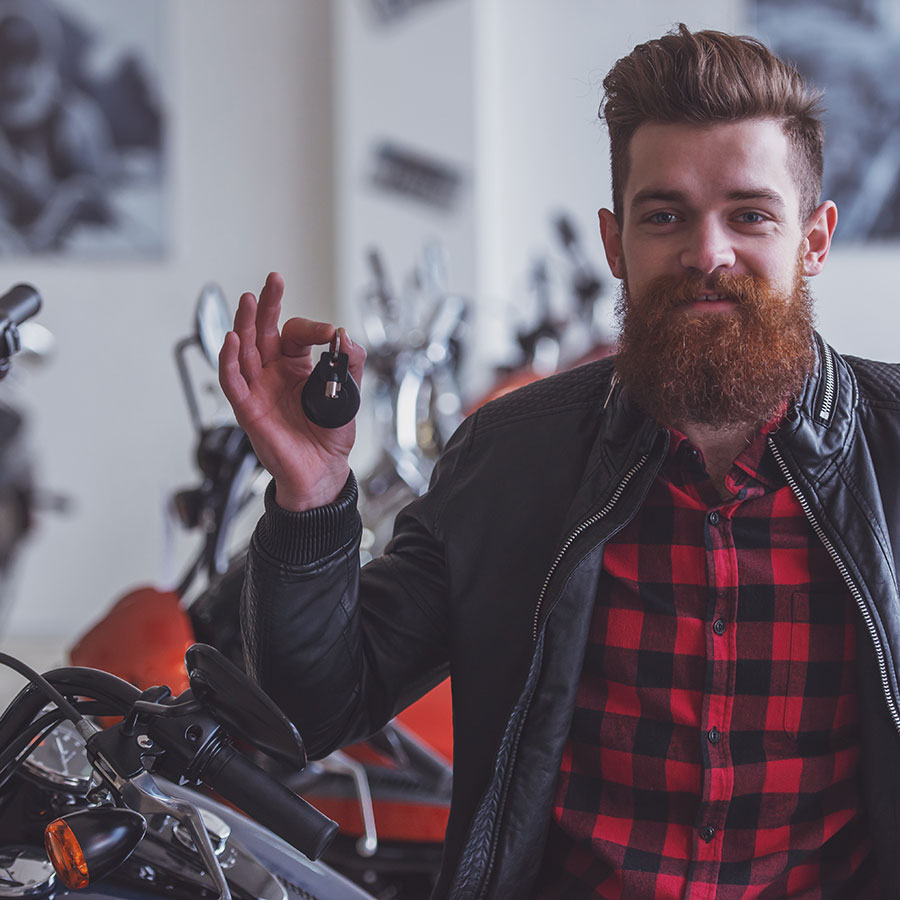 A man in a plaid shird with a black leather jacket over top is holding a key to a newly financed adventure vehicle. He has a big smile on his face and a brown, bushy beard.