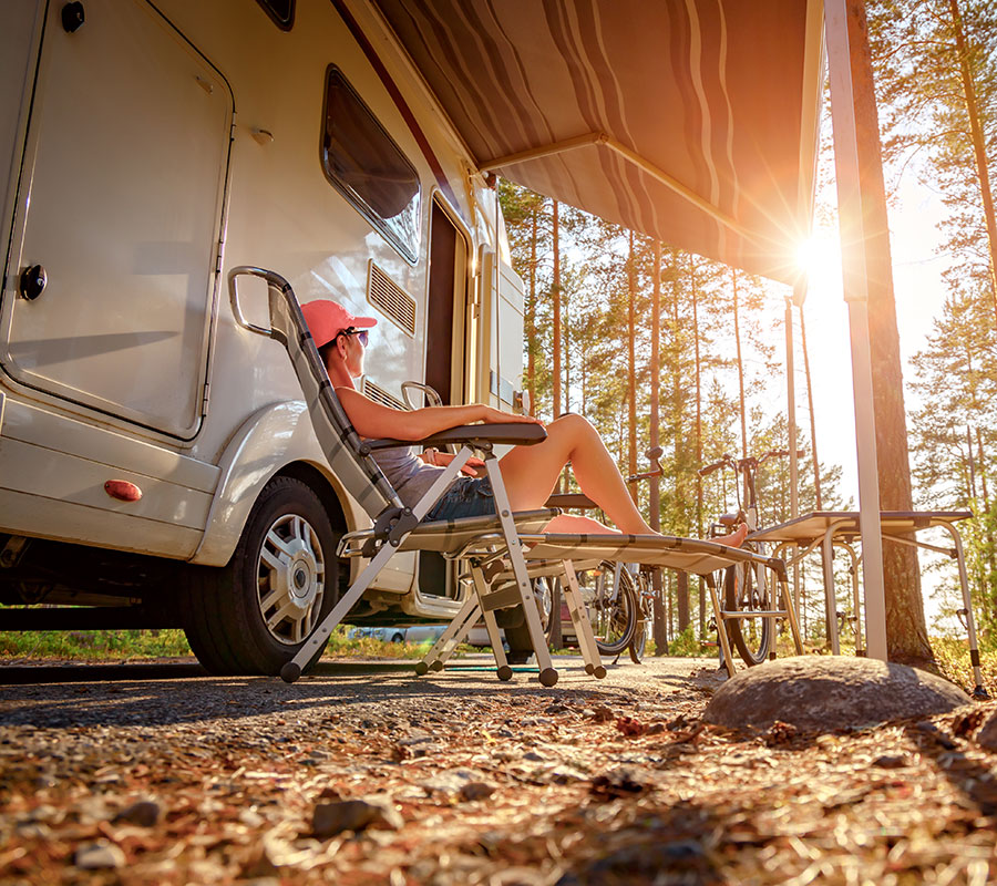 A white woman with a pink ballcamp relaxes on a camp chair in the late afternoon sun. She has her feet up and is under an awning alongsider her newly financed RV.