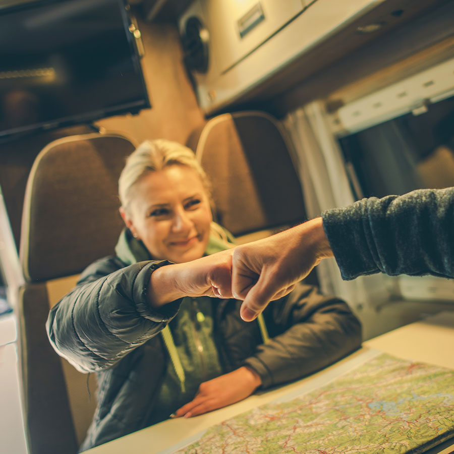 A woman fist bumbs a man in a RV. They are sitting at the kitchenette and are reviewing a map on the kitchen table, prepping for adventure.